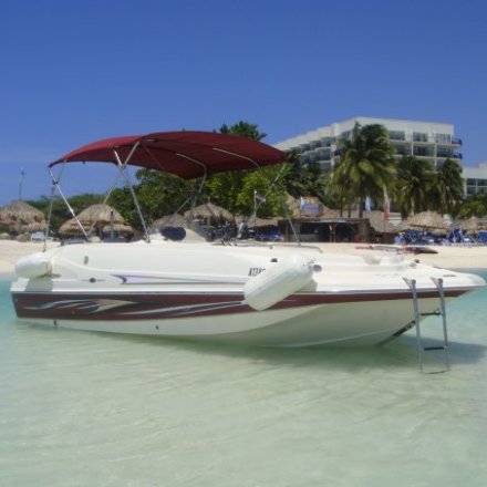 A small yacht with maroon cover & stripe on the side sitting in shall water with cabanas, palm trees and a hotel in the background